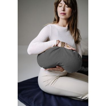 Kobieta w jasnym stroju trzyma szarą poduszkę do medytacji w kształcie fasolki na dłoniach. W tle leżą ułożone poduszki i mata do medytacji.